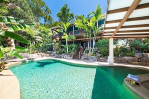 A clear pool surrounded by tropical vegetation and a multi-level building with a wooden deck.