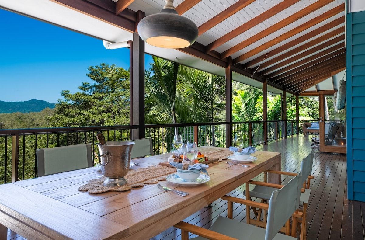 A wooden dining table set with plates, cutlery, and a pitcher, under a hanging light fixture, with a view of greenery and mountains.