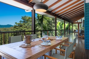 A wooden dining table set with plates, cutlery, and a pitcher, under a hanging light fixture, with a view of greenery and mountains. - Casa Mariposa—A Secluded Tropical Sanctuary (Riordanvale)