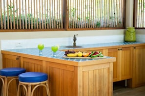 A kitchen with a wooden island, two stools, a fruit display, and a bamboo screen. - Casa Mariposa—A Secluded Tropical Sanctuary (Riordanvale)