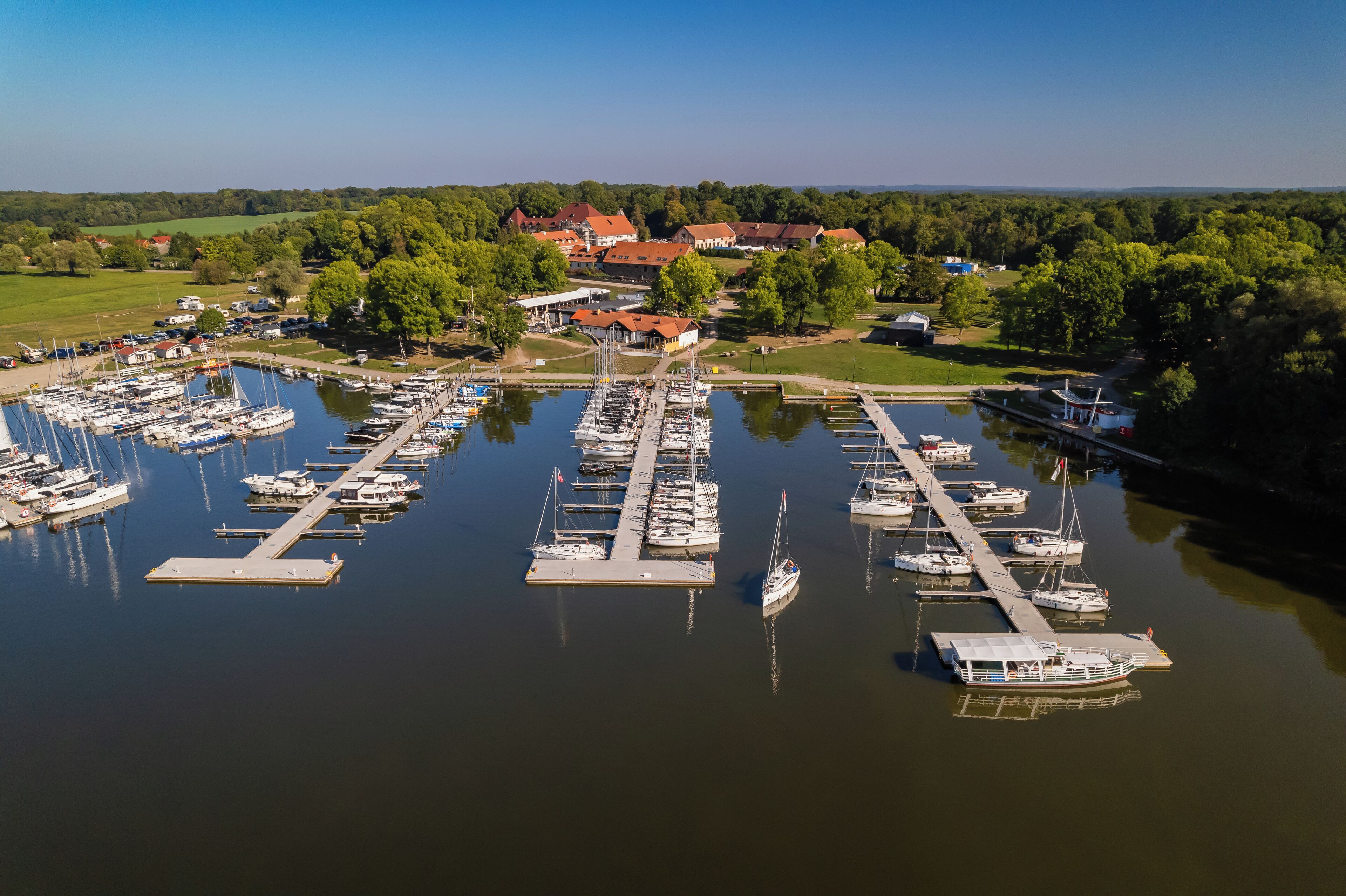 A marina with numerous boats docked at piers, surrounded by greenery and buildings in the background.