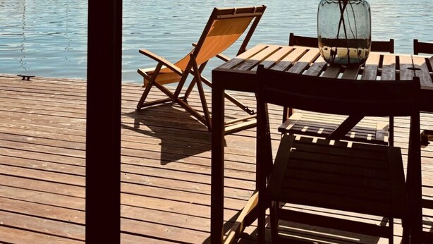 Een houten terras met een tafel en stoelen, uitkijkend op een rustig meer met bootjes en een heldere lucht.