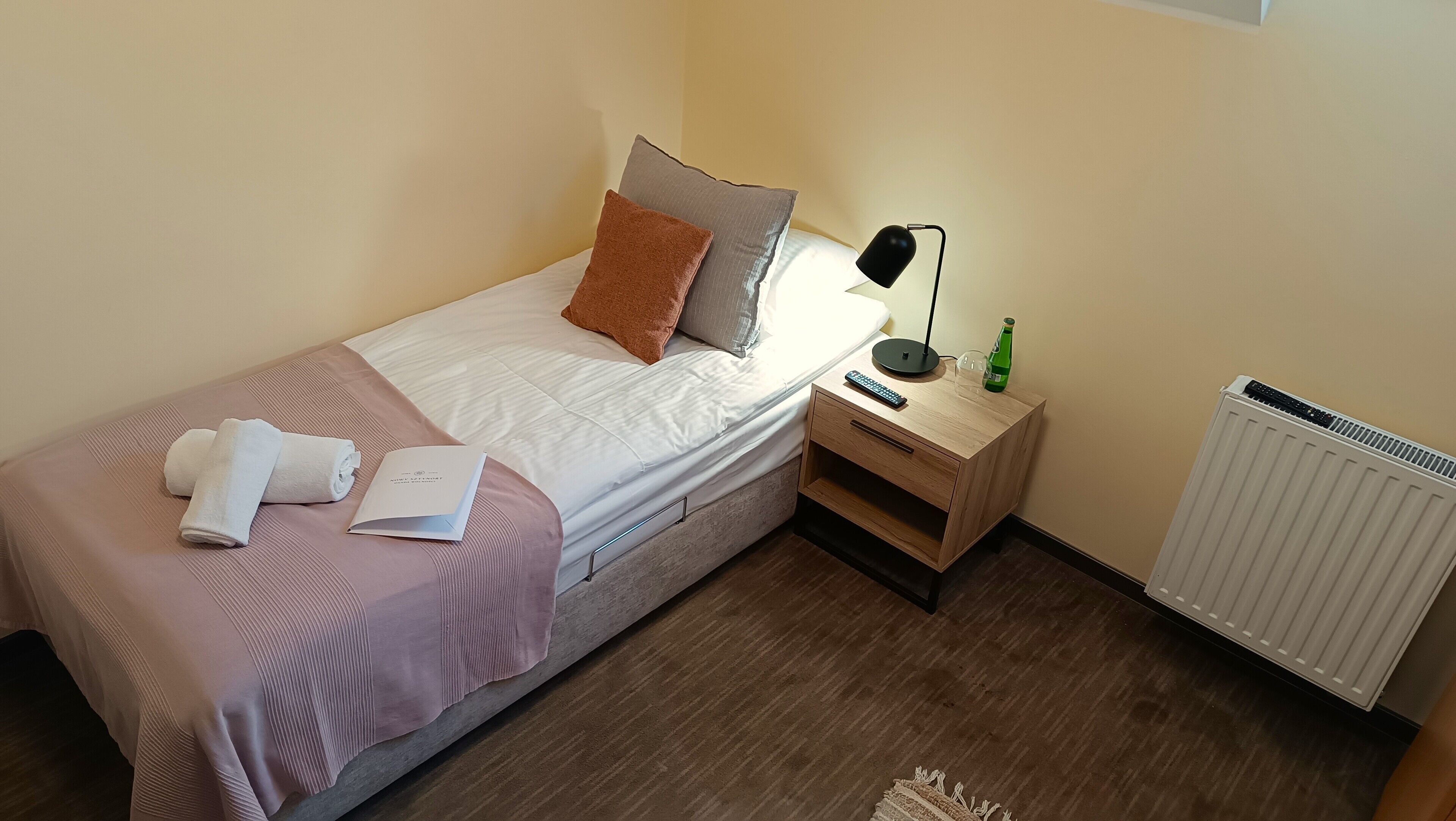 Un letto rifatto con una coperta bianca e rosa, un telecomando, una bottiglia e un libro su un comodino di legno.