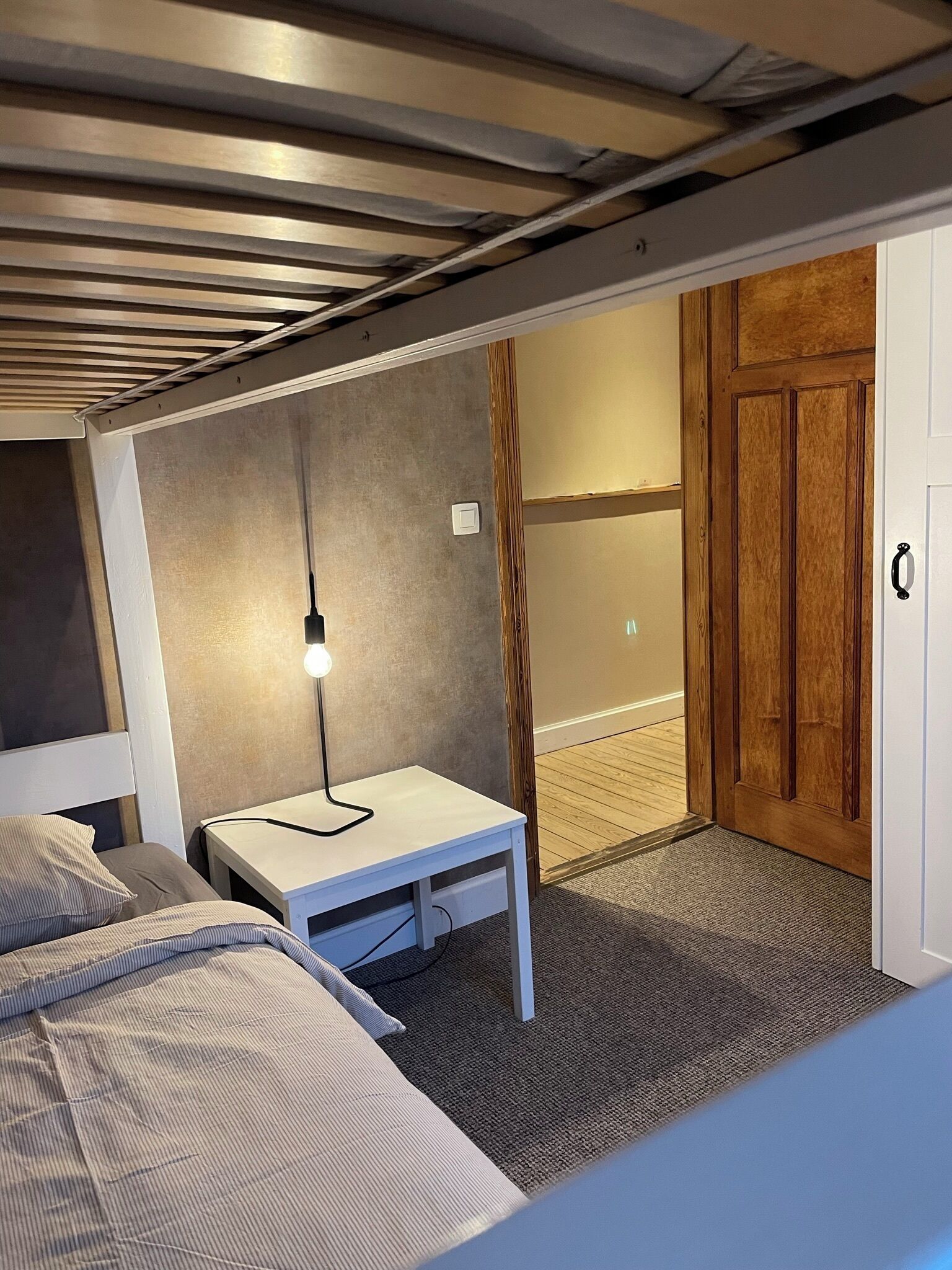 A compact room with a bed, a small table with a lamp, a wooden door, and a textured wall.