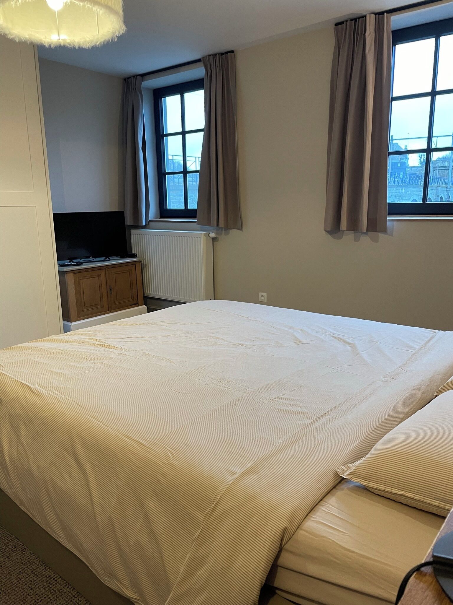 Ein Schlafzimmer mit einem großen Bett, einem Heizkörper, einem Fernseher auf einer Holzkommode und zwei mit Vorhängen versehenen Fenstern.