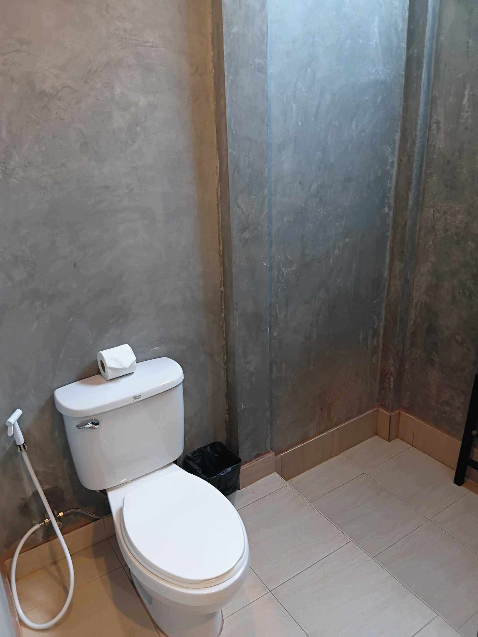 Ein Badezimmer mit Toilette, einer Toilettenrolle, einer schwarzen Mülltonne und einem weißen Schlauch.