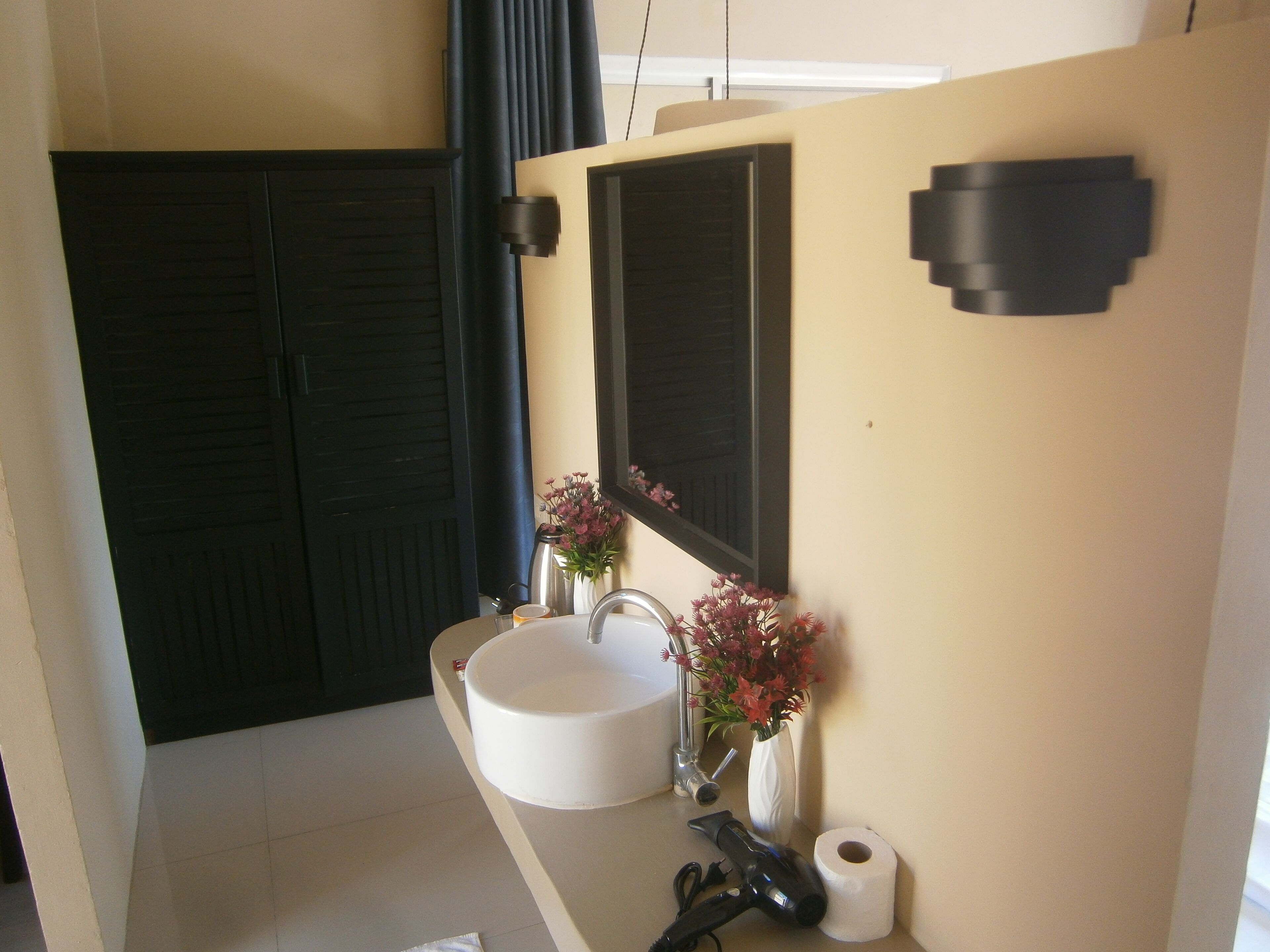 A bathroom with a white sink, a mirror, a vase with flowers, and a toilet paper roll.