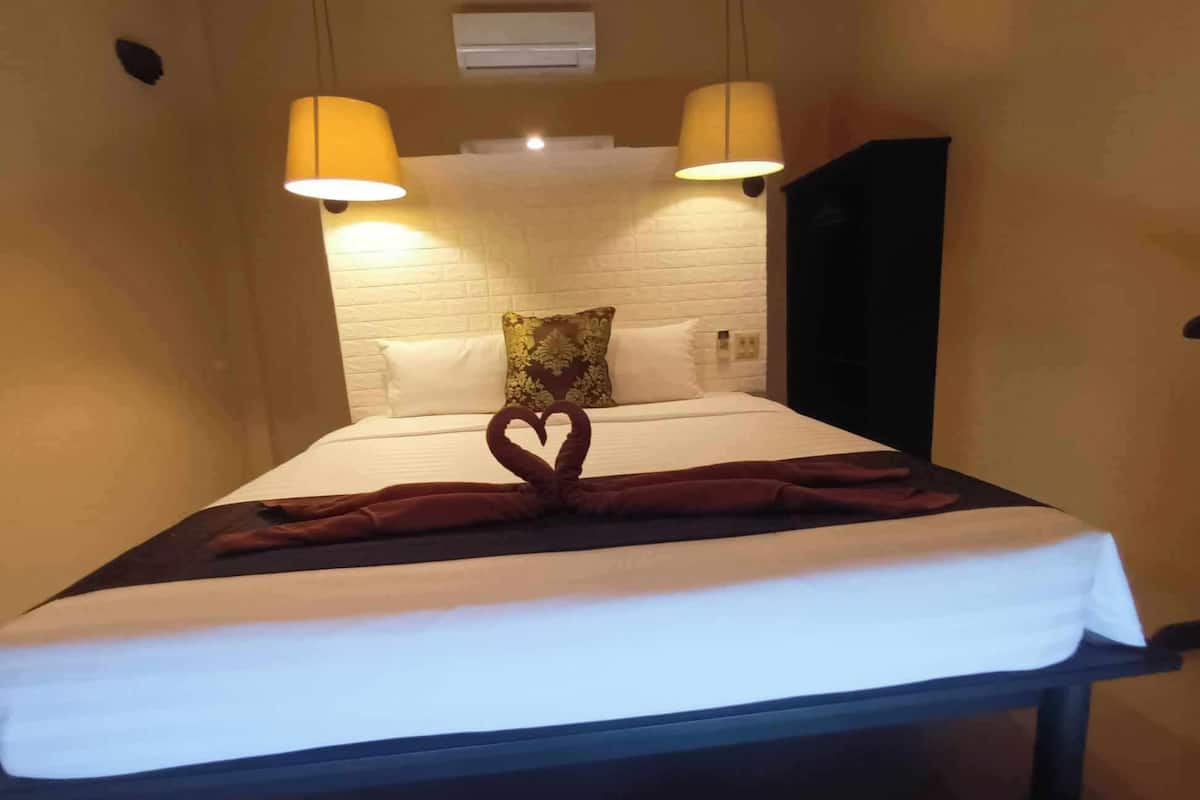Habitación de hotel con una cama bien hecha, dos lámparas de pared, cabecera de ladrillo blanco y una toalla doblada en forma de corazón.