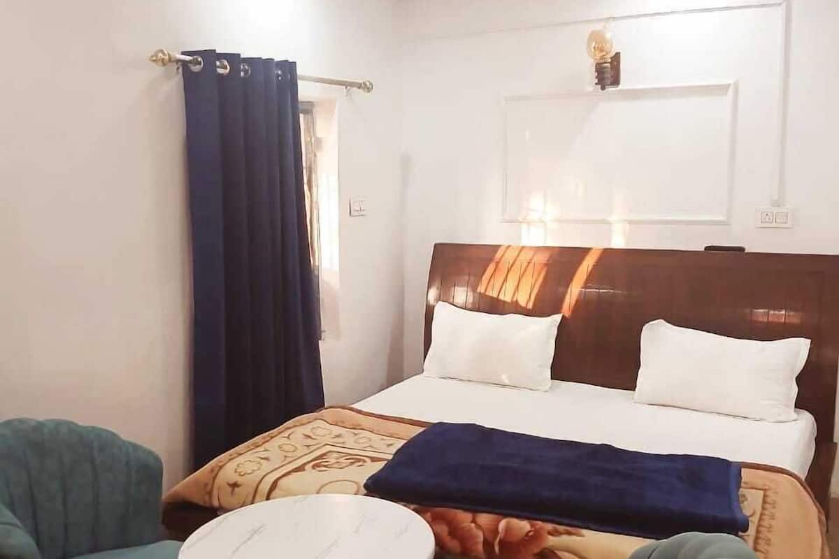 Um quarto com cama, duas cadeiras, uma mesinha, ventoinha no teto e janela com cortinas.
