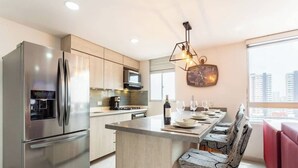 A modern kitchen with a stainless steel refrigerator, microwave, and oven. There is a dining area with a table set for four and a bar stool. - Luxe Apartment: By Medellín, Park, Dining, Nightlife, Fast WiFi & Self Check-in (Sabaneta)
