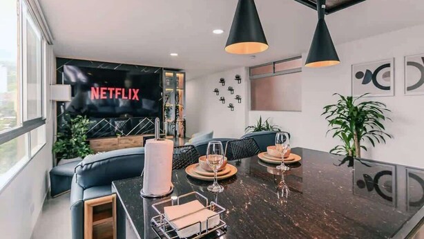 A modern living room with a dark wooden dining table, black pendant lights, a large flat-screen TV, and a cozy seating area with a sofa and cushions.