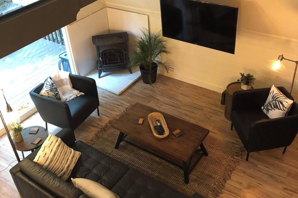 A modern living room with a black leather sofa, two armchairs, a wooden coffee table, and a flat-screen TV mounted on the wall.