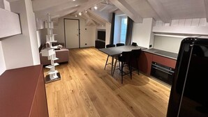 Une pièce aménagée en style mansardé, avec un plancher en bois, une table à manger et des chaises, un coin cuisine avec une cuisinière, et un escalier agrémenté d’une étagère blanche.