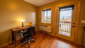 Ein Zimmer mit Schreibtisch aus Holz, Stuhl, Lampe und Fenster mit Blick auf eine Terrasse und Bäume.