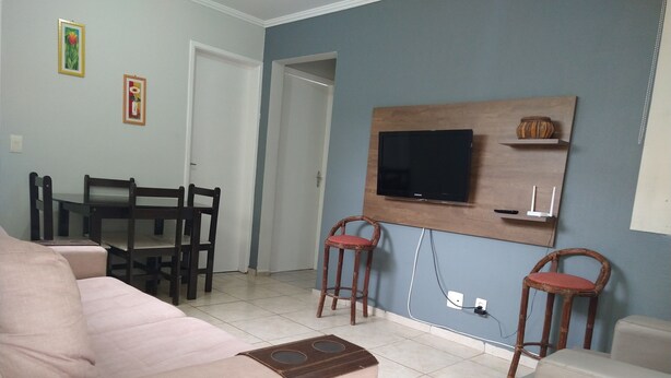 A living room with a sofa, two chairs, a TV mounted on the wall, and a dining table with chairs. - Round trip - LA04-Bertioga, SESC, Riviera, Vista Linda, Indaiá  (Jardim Rafael Bertioga)