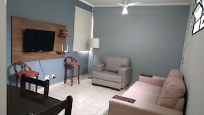 A living room with a television mounted on a wooden panel, a ceiling fan, a sofa, a chair, and a table. - Round trip - LA04-Bertioga, SESC, Riviera, Vista Linda, Indaiá  (Jardim Rafael Bertioga)