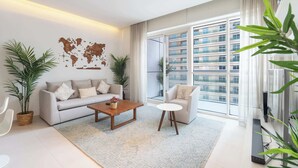A modern living room with a sofa, a coffee table, and a small side table. There are two potted plants, a world map on the wall, and a large window with a view of a high-rise building.