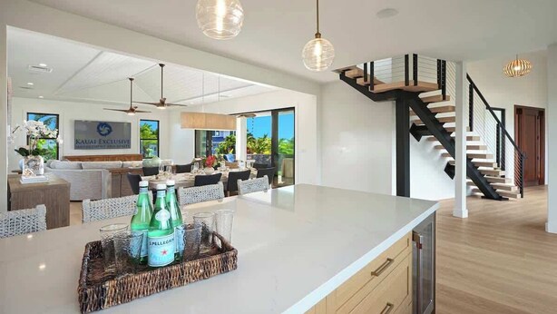 A modern kitchen with a large island, a dining area with a sofa and table, and a staircase with a black railing.