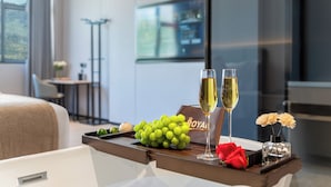Ett hotellrum med en säng, ett nattduksbord med ett bricka som innehåller druvor, två glas champagne, en röd ros och en liten vas med blommor.