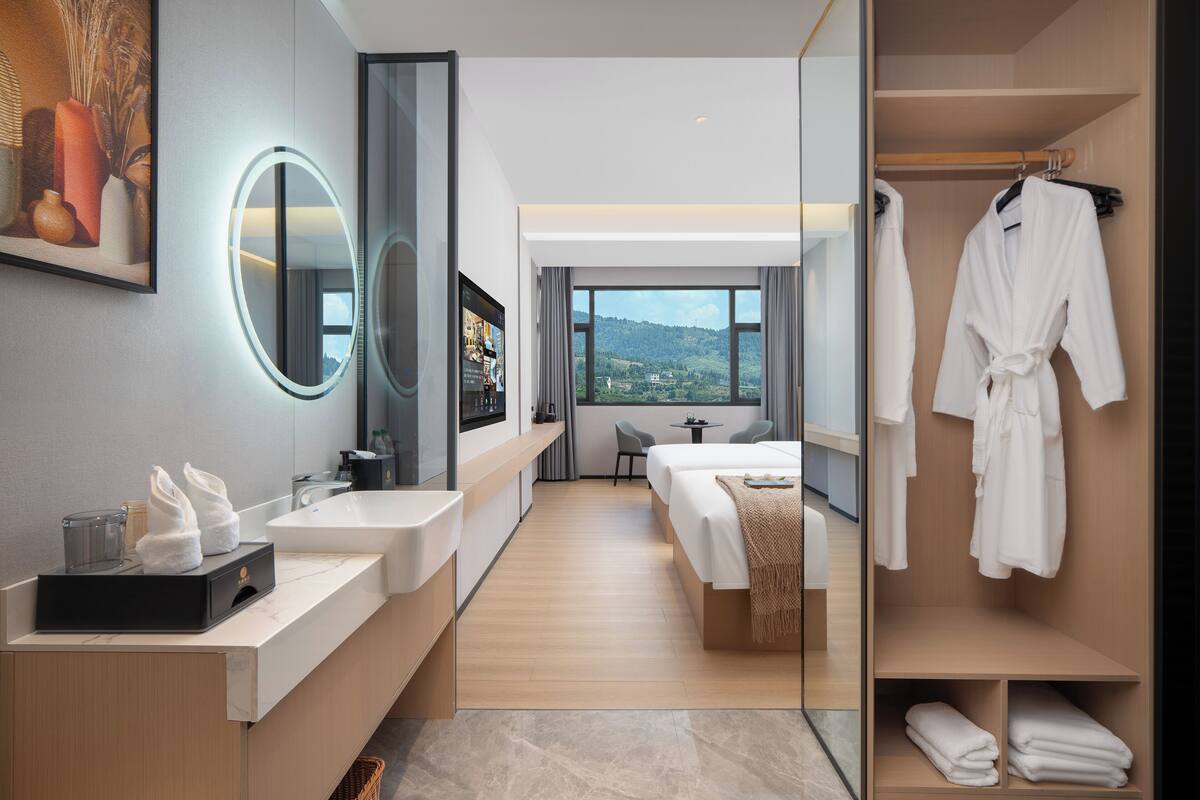 A modern hotel room with a large mirror, a vanity with a sink, a bed, a bedside table, and a view of the outdoors.