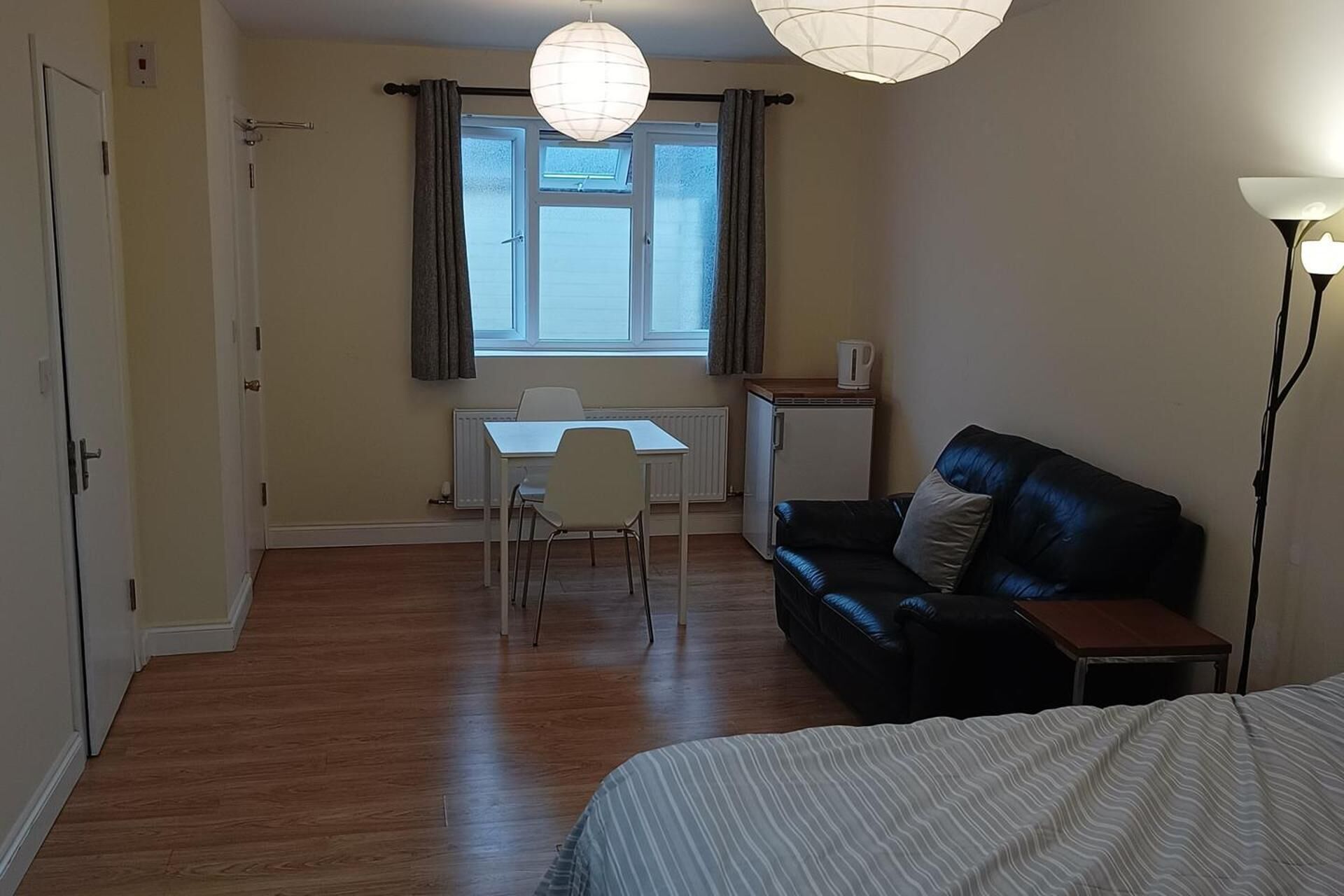 A small, well-lit room with a wooden floor, a black leather sofa, a white desk with a chair, a radiator, and a window with curtains.