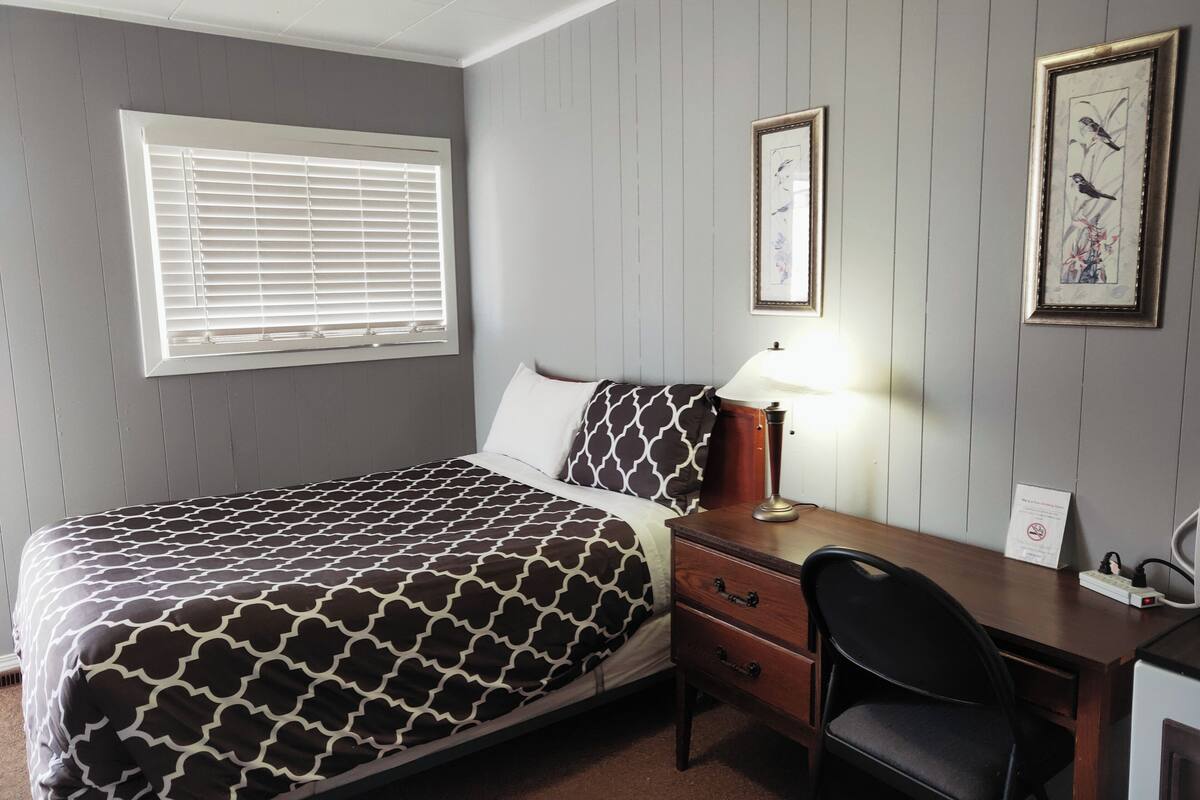 A room with a bed, a desk, a chair, a lamp, and two framed pictures on the wall.