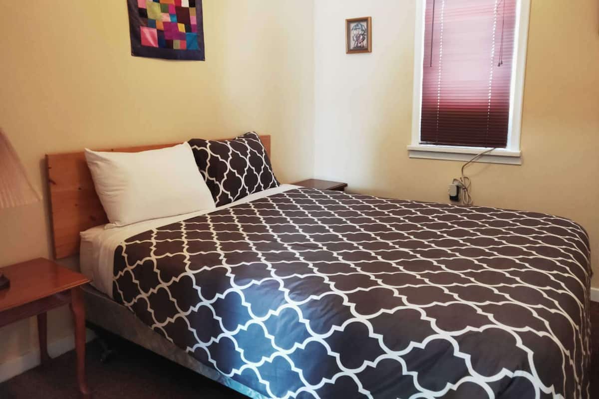 A neatly made bed with a patterned bedspread, a bedside table, a small framed picture, and a window with blinds.