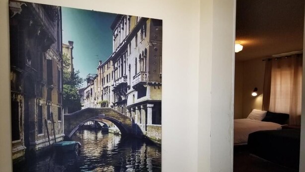 A hotel room with a large painting of a canal and bridge, a bed, and a bedside lamp.