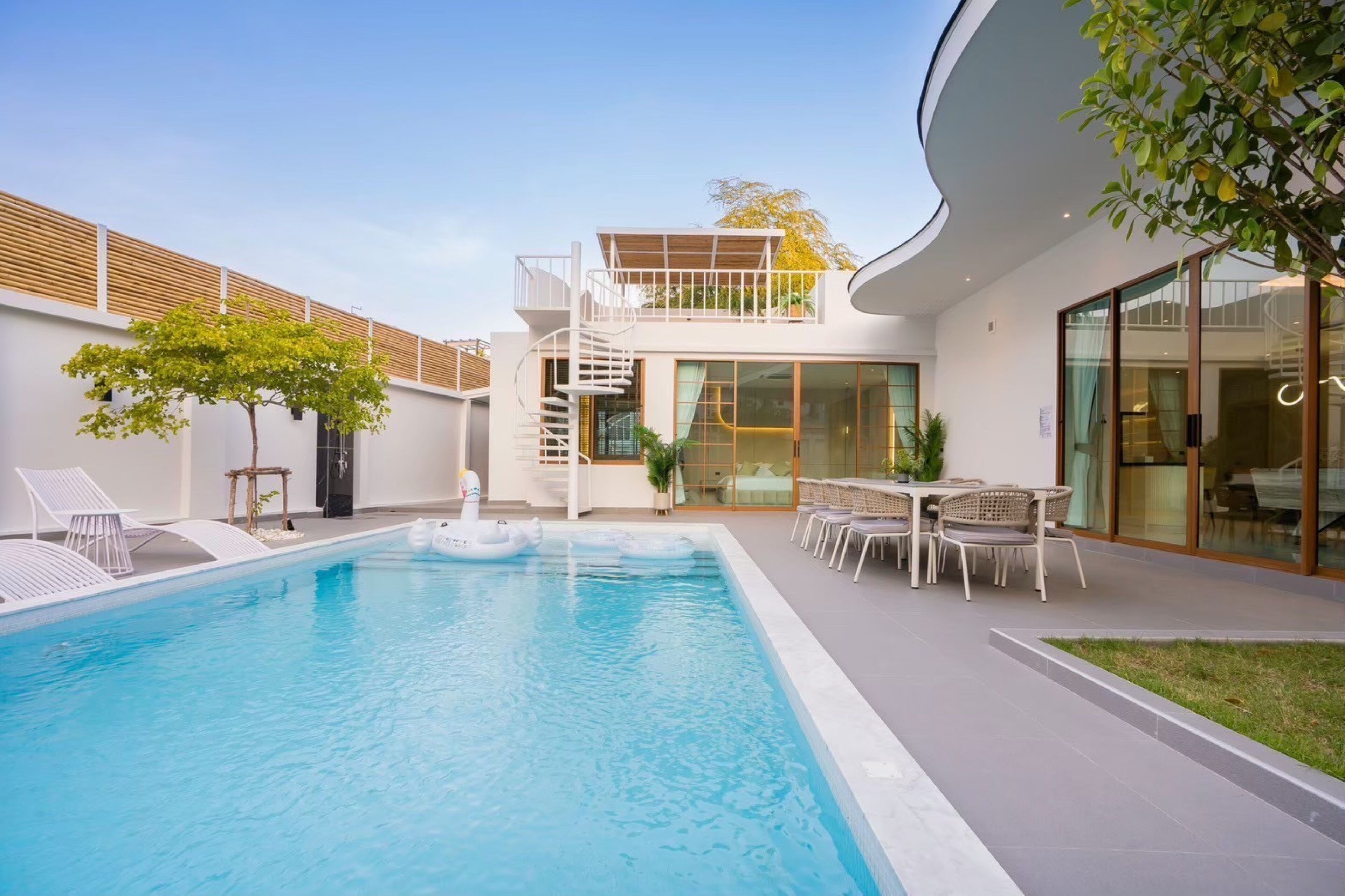 Een modern zwembadgedeelte met een helderblauw zwembad, ligbedden, een eethoek met tuinmeubilair en een trap naar een balkon op de tweede verdieping.