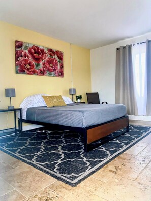 Une chambre à coucher comprenant un lit, deux tables de chevet, une lampe, un grand tableau représentant des fleurs et une fenêtre avec des rideaux.