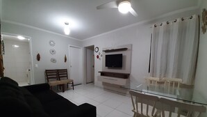A living room with a glass dining table, wooden chairs, a black sofa, a TV mounted on the wall, and a ceiling fan. - Round Trip - LA05-Bertioga, SESC, Riviera, Vista Linda, Indaiá (Jardim Rafael Bertioga)