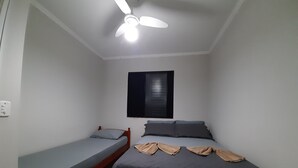 A small, single-room accommodation with a bed, a single bedside table, a ceiling fan, and a window with blinds. - Round Trip - LA05-Bertioga, SESC, Riviera, Vista Linda, Indaiá (Jardim Rafael Bertioga)