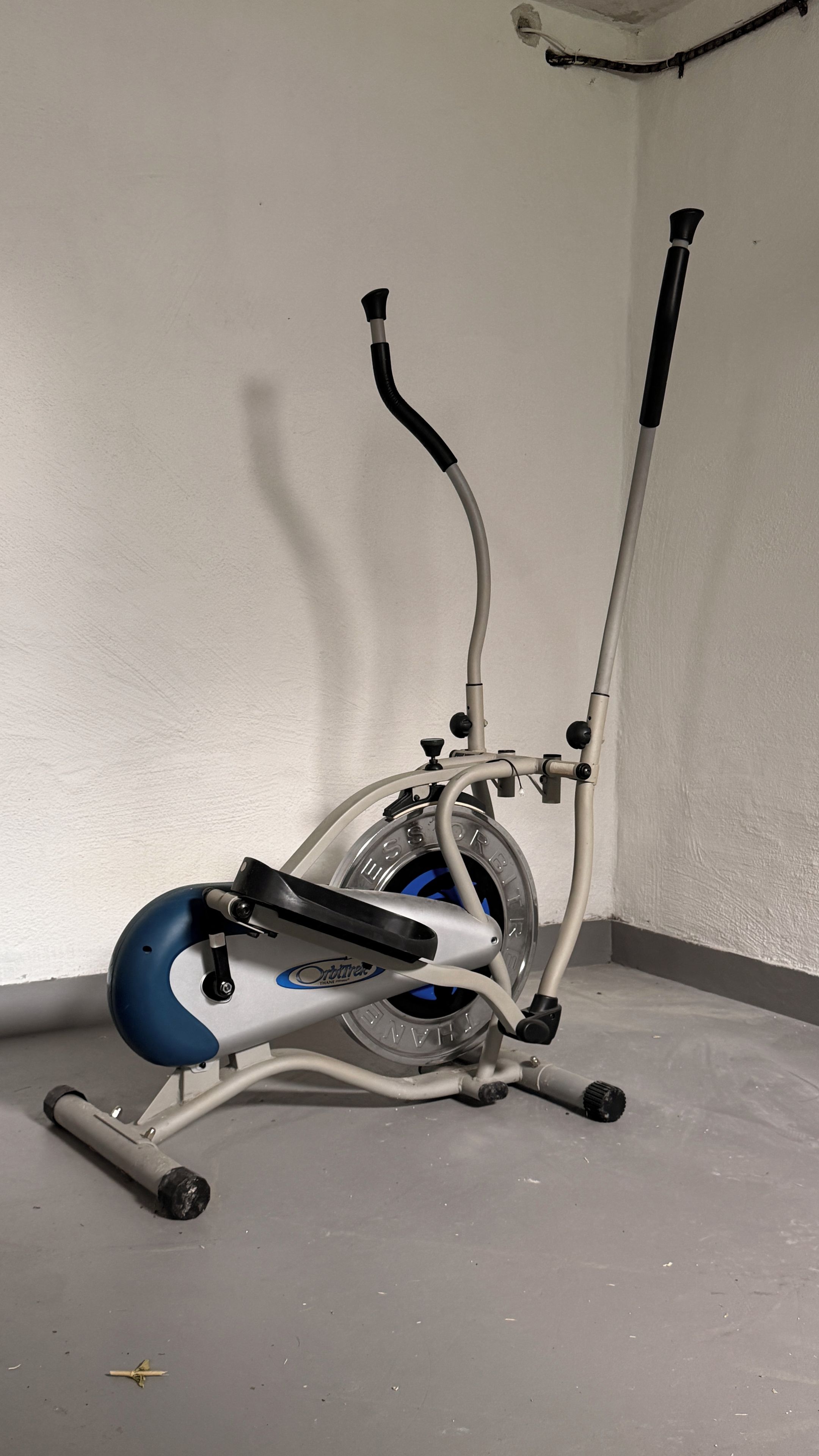 An exercise bike with a blue seat and handlebars, positioned in a corner of a room with a plain wall and a concrete floor.