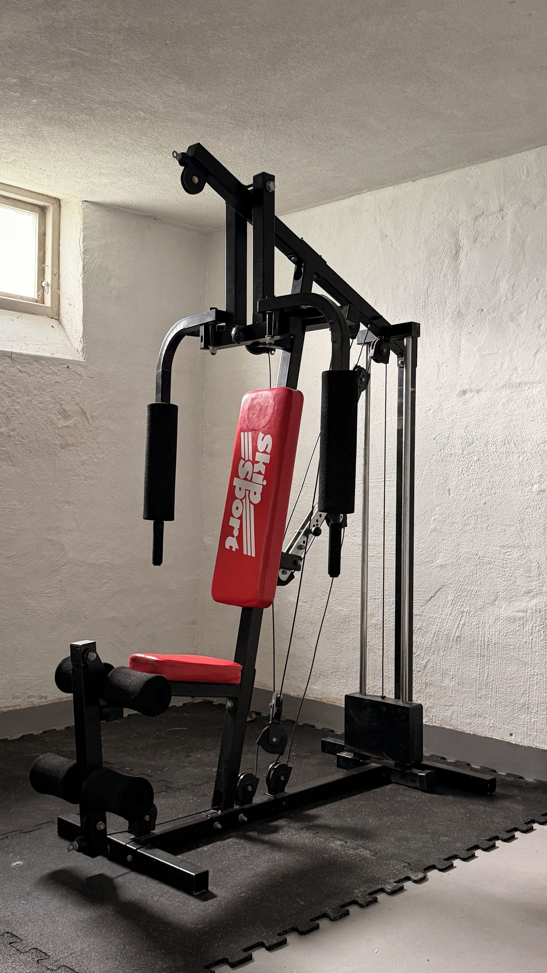 Une salle de sport avec un banc rouge, des appareils de musculation et un système de poulies fixé au mur.