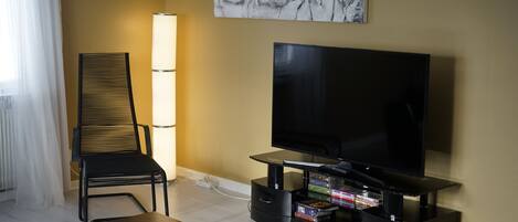A modern living room with a flat-screen TV, a black chair, a table with a remote control, and a wall-mounted artwork.