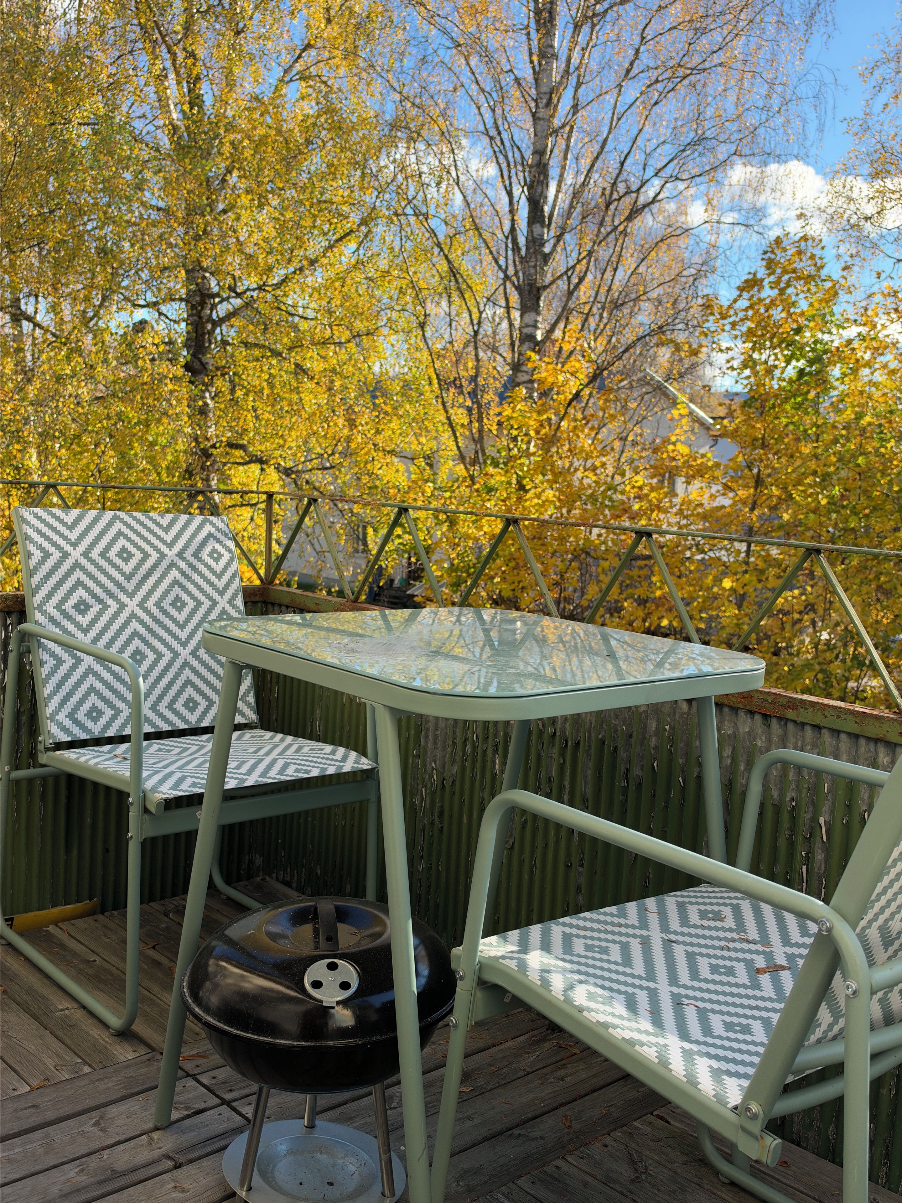 A patio with two chairs and a table, surrounded by a wooden deck and trees with yellow leaves.