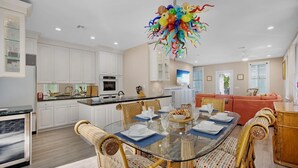 Une cuisine moderne avec des meubles blancs, un îlot central et un coin repas comprenant une table en verre et des chaises en osier.