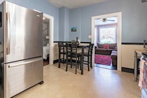 A modern kitchen with a stainless steel refrigerator, a dining area with dark wooden chairs, and a view into a living room with a brown sofa and a ceiling fan.