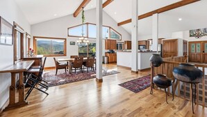 A spacious kitchen with wooden cabinets, a dining area with a table and chairs, and large windows offering a view of the outdoors. - Gold Dust Getaway | Hot tub | Near Ski Resorts (Edwards)
