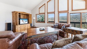 A spacious living room with a large wooden entertainment center, leather sofas, a patterned rug, and large windows offering a view of the outdoors. - Gold Dust Getaway | Hot tub | Near Ski Resorts (Edwards)