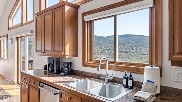 A kitchen with wooden cabinets, a stainless steel sink, a coffee maker, and a view of a landscape through large windows. - Gold Dust Getaway | Hot tub | Near Ski Resorts (Edwards)