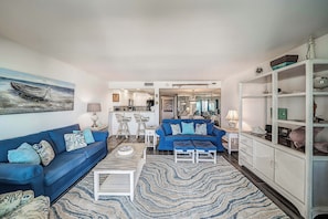 A living room with a blue sofa, a coffee table, and a patterned rug. There is a kitchen in the background with white cabinets and a dining area with a blue table. - FREE Golf & Perks! Mariner West 504 Beachfront Swimming Pool, Tennis & Grill! (Panama City Beach)