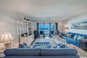 A living room with a blue sofa, a white shelving unit, and a view of the ocean through large windows. - FREE Golf & Perks! Mariner West 504 Beachfront Swimming Pool, Tennis & Grill! (Panama City Beach)