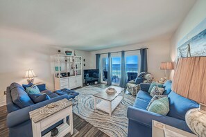 A modern living room with a blue sofa set, a white entertainment unit, and a large window with a view of the ocean. - FREE Golf & Perks! Mariner West 504 Beachfront Swimming Pool, Tennis & Grill! (Panama City Beach)