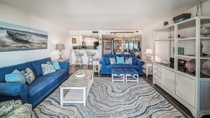 A living room with a blue sofa, a coffee table, and a patterned rug. There is a kitchen in the background with white cabinets and a dining area with a blue table.