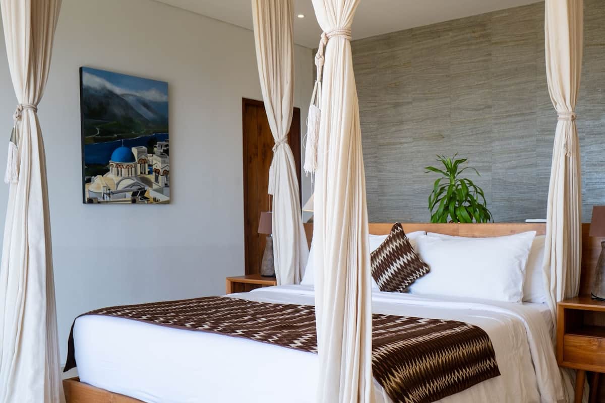 A bedroom with a four-poster bed, a patterned rug, a painting on the wall, and a plant in the corner.