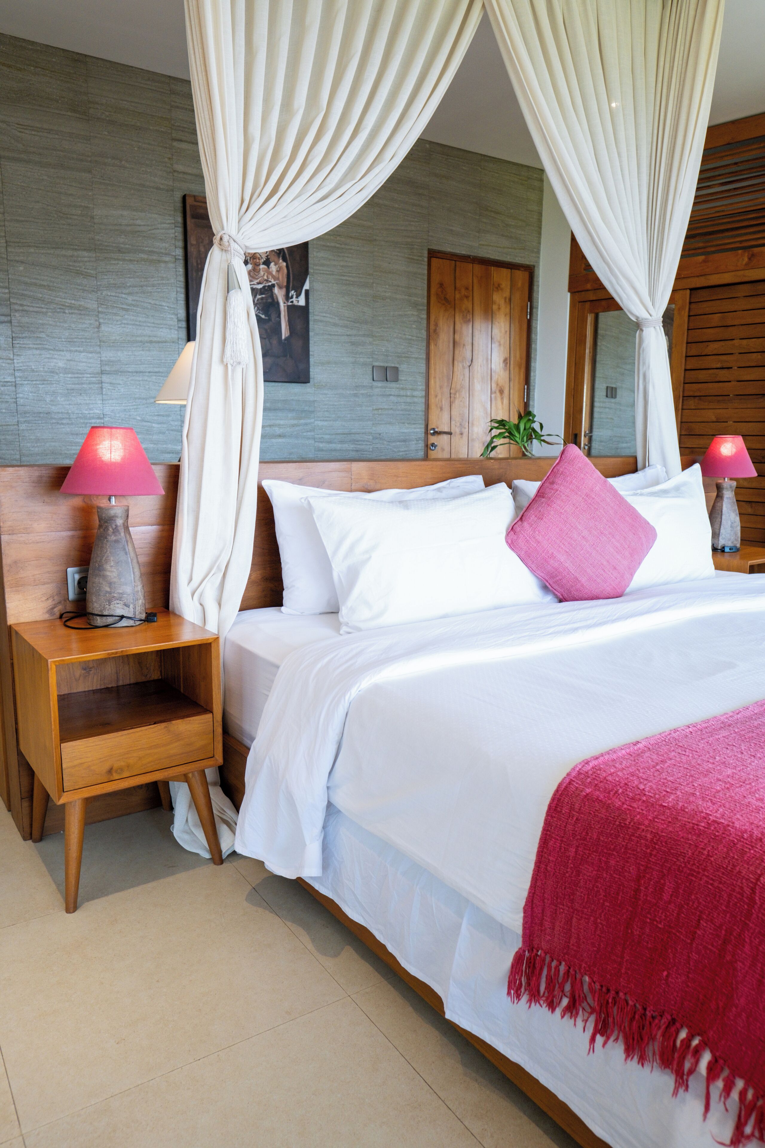 A neatly made bed with white bedding and a pink throw, a wooden nightstand with a lamp, and a large window with curtains.