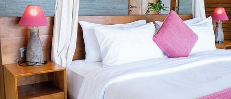 A neatly made bed with white bedding and a pink throw, a wooden nightstand with a lamp, and a large window with curtains.
