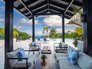 A covered outdoor seating area with white sofas and chairs, a wooden table, and a pool in the background. - 13 bedroom Ultimate Private Luxury Compound with every amenity you can imagine (Sosúa)