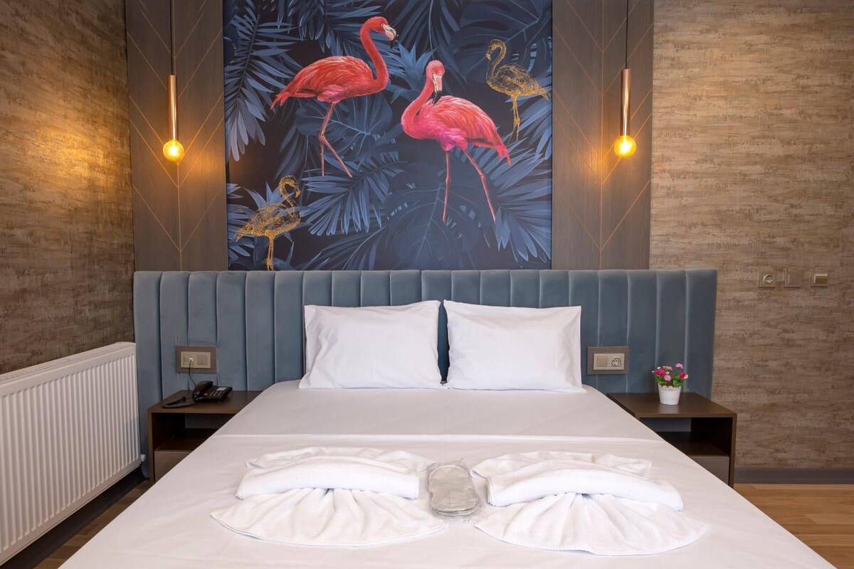A hotel room with a large bed, two bedside tables, a nightstand, a wall-mounted light, and a tropical-themed painting.